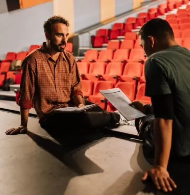 Dos actores sentados en el borde del escenario revisan sus guiones en un teatro con butacas rojas, durante una sesión de coaching para actores de Terapia Escénica.