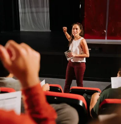 Paula Sanz habla a un grupo de alumnos en un teatro con butacas rojas durante una sesión para escuelas de Arte Dramático (Terapia Escénica).